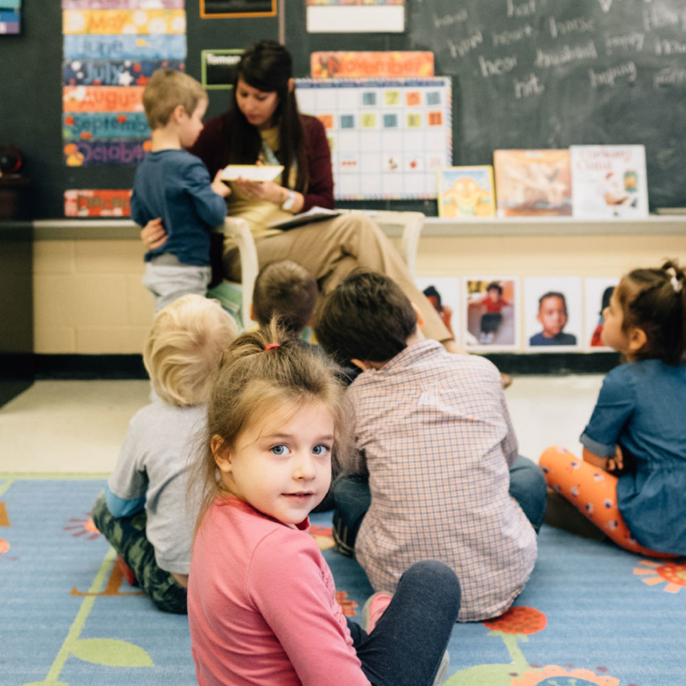 Preschool classroom with children sitting on the floor listening to a teacher